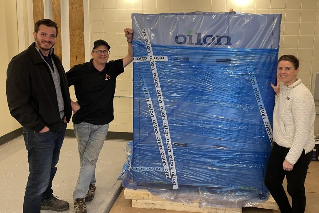 A newly delivered Oilon ChillHeat heat pump, still wrapped in blue plastic and Oilon-branded tape on a wooden pallet, in a basement mechanical room ready for installation.