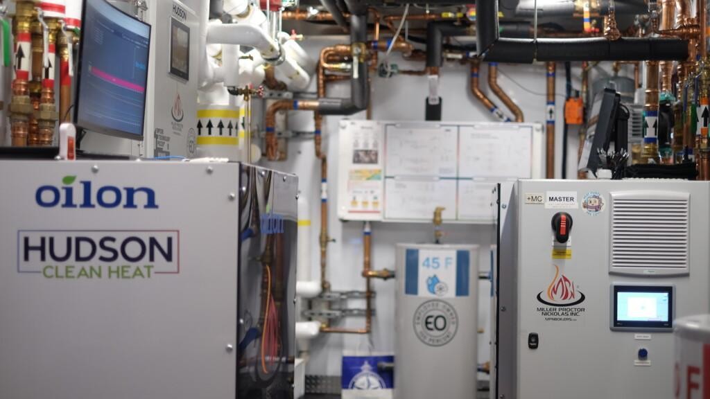 Inside the award-winning mobile demonstration trailer: an Oilon heat pump with Hudson Clean Heat branding beside a Miller Proctor Nickolas control unit, surrounded by copper piping, showing how compact the steam retrofit solution is.