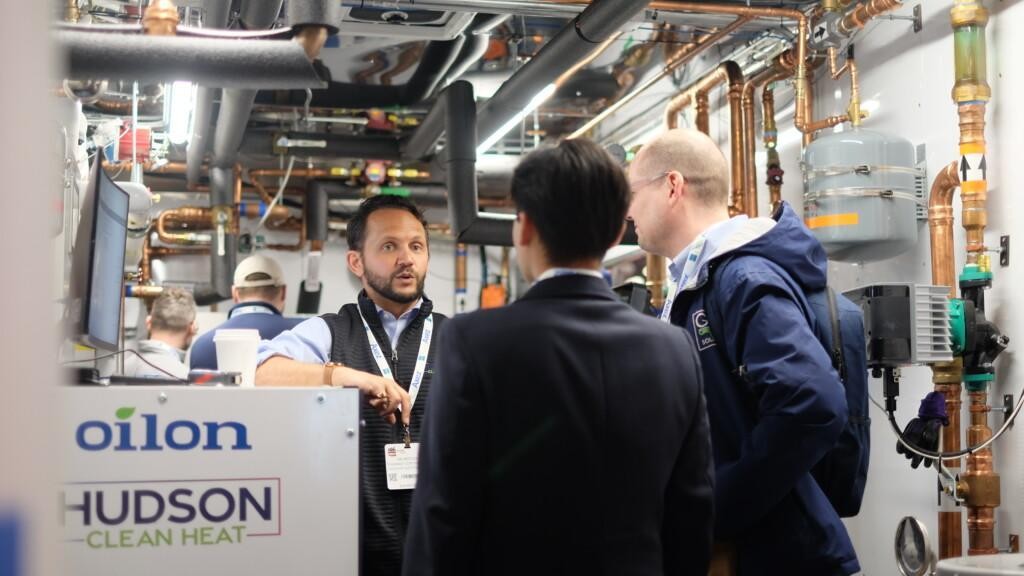 Ian Motley, President of Hudson Clean Heat, explains the Oilon and Hudson Clean Heat retrofit system to visitors inside the mobile demonstration trailer, standing behind an Oilon heat pump among copper piping.